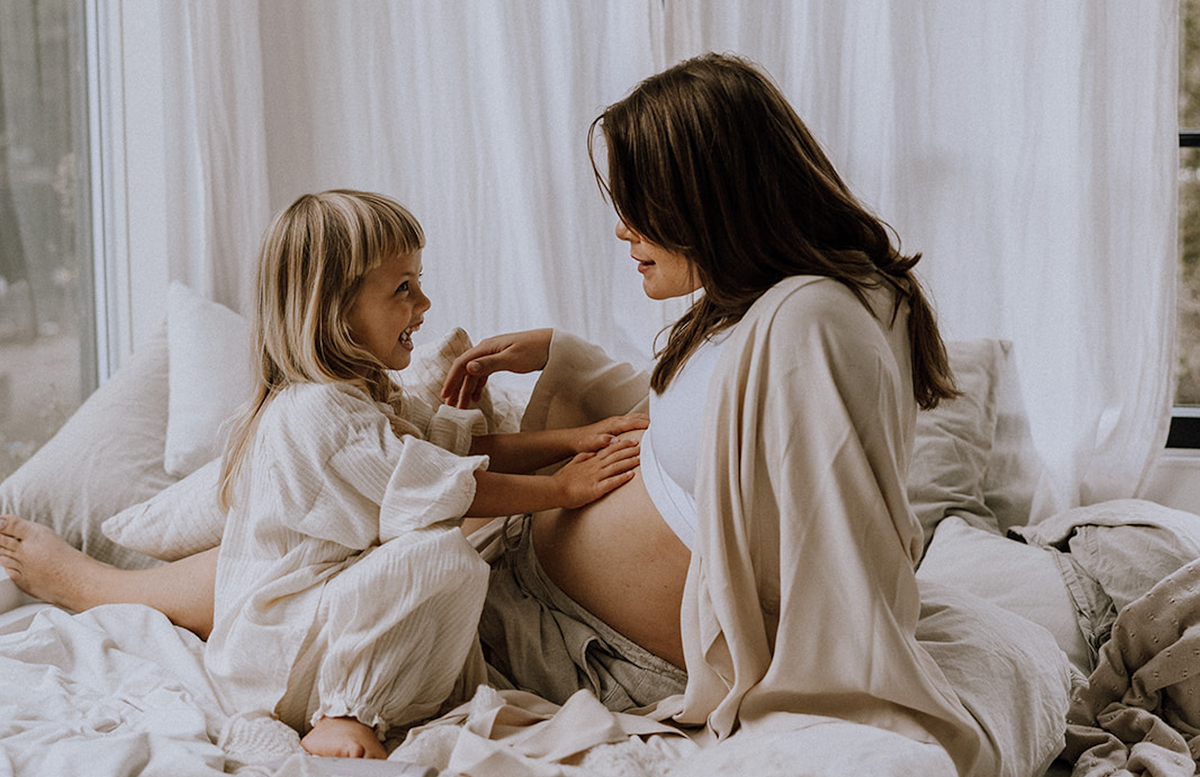 Gravid kvinde sidder på sengen, mens et barn smilende lægger hånden på hendes mave.